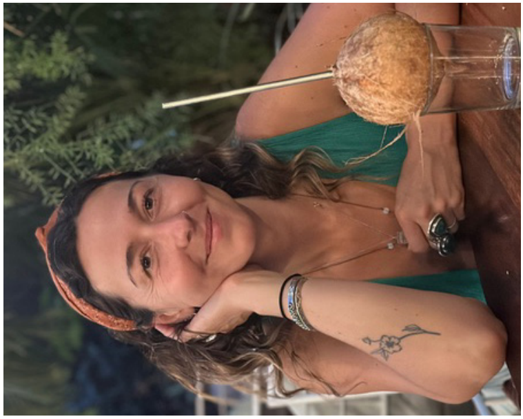 Portrait of Emery Jane Sunderland Golson smiling warmly and resting her chin on her hand, with a coconut beverage beside her