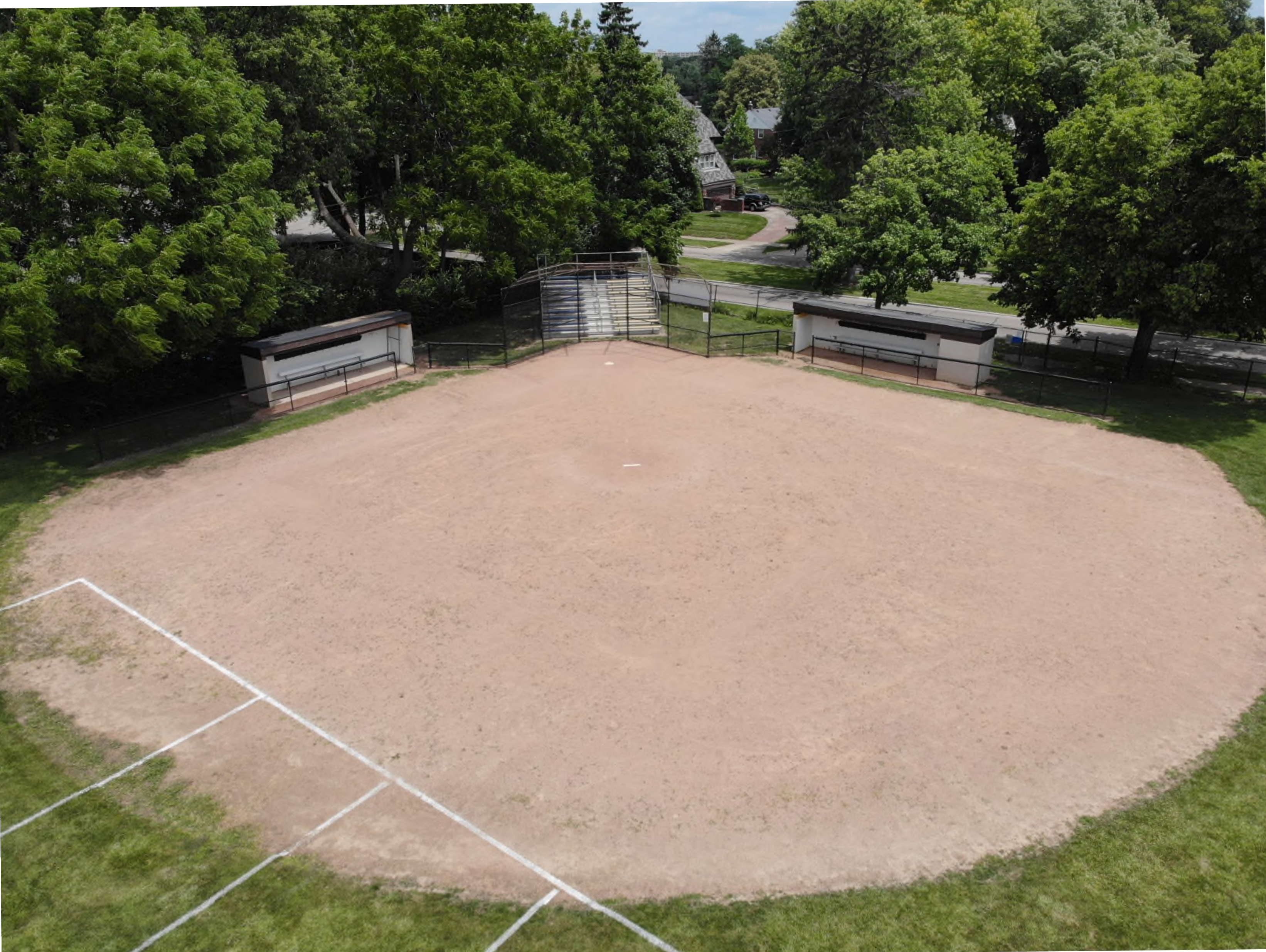 Aerial view of a baseball and softball dirt infield featuring the pitcher's mound, home plate, backstop fencing, player dugouts, bleachers, and surrounding green turf and trees.