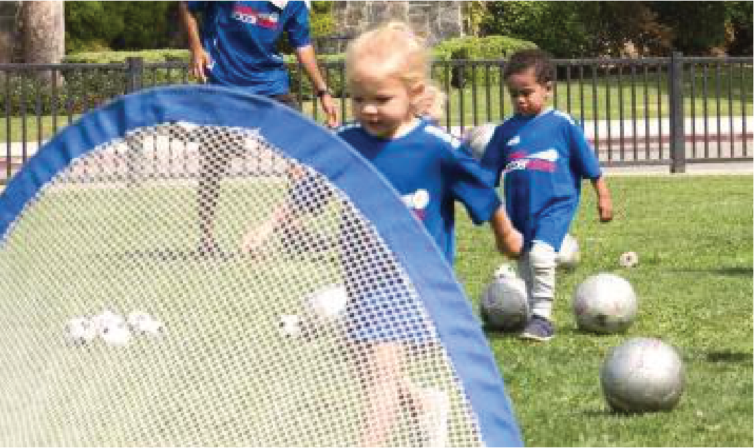 Two young children in blue soccer jerseys playing with soccer balls on a grass field behind a portable goal net.