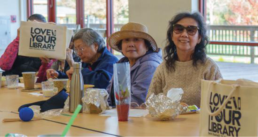Two older women at a community center table with 'Love Your Library' tote bags