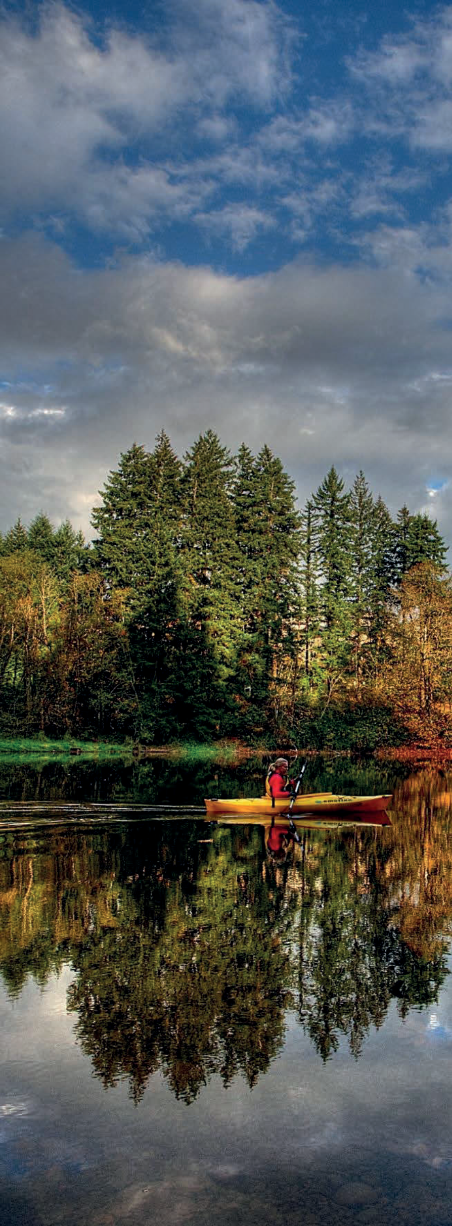 Kayaker paddling on a scenic lake reflecting evergreen trees under a cloudy sky