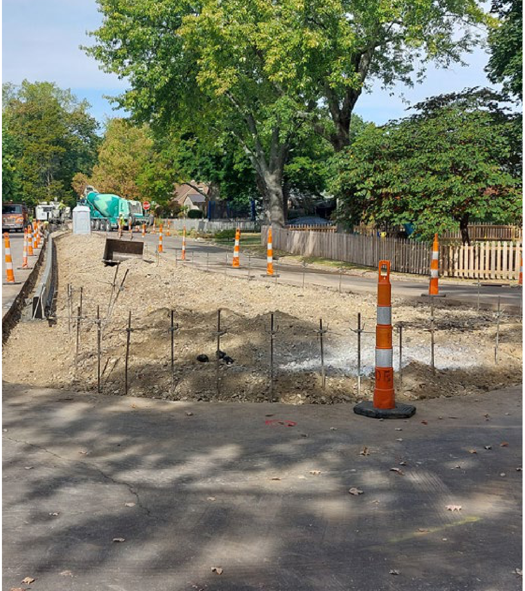 Construction phase of Harman Boulevard median showing excavation, stakes, safety barrels, and a concrete truck in the background