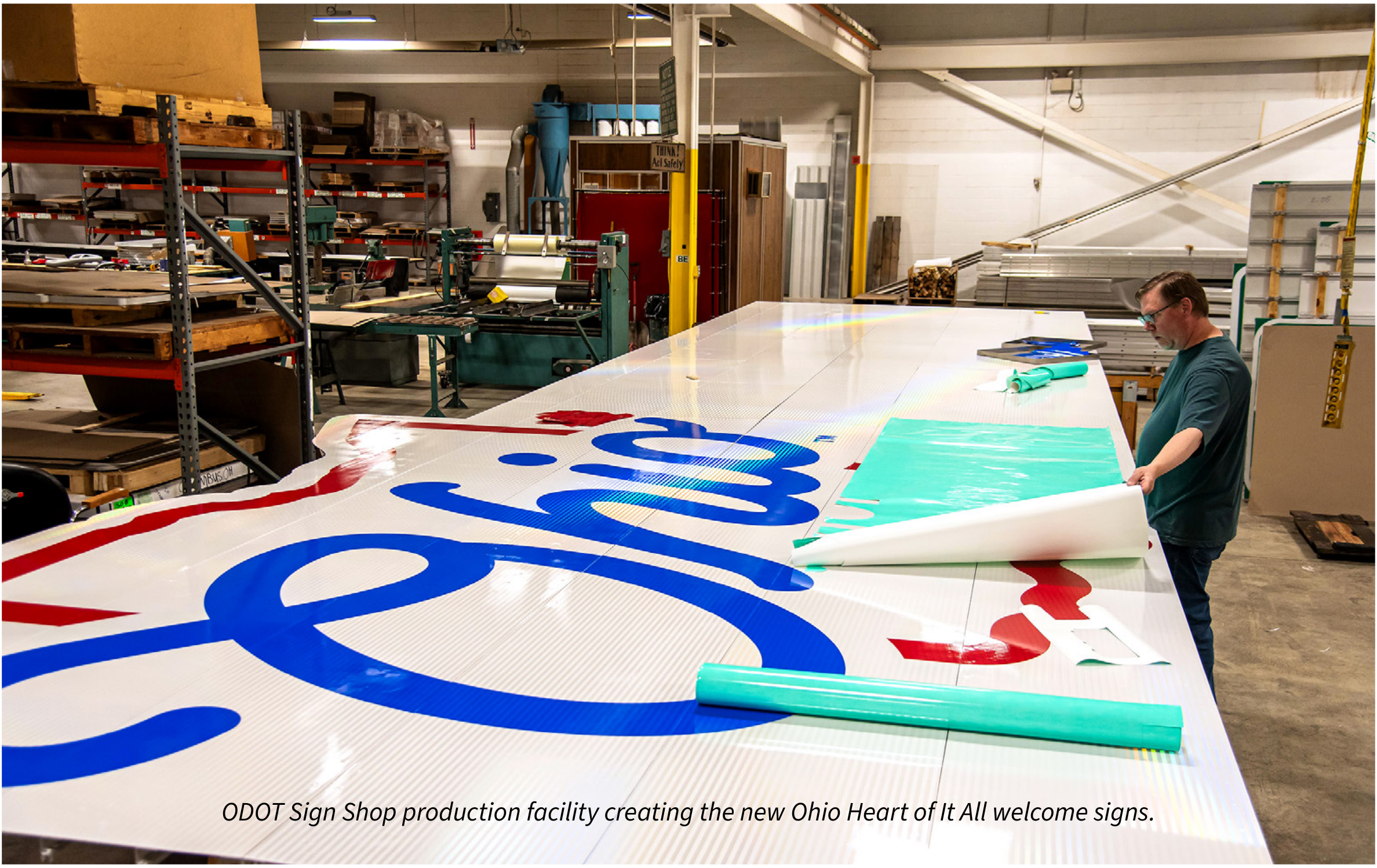A worker at the ODOT Sign Shop applying vinyl sheeting to construct a large 'Ohio The Heart of It All' highway welcome sign in a production warehouse.