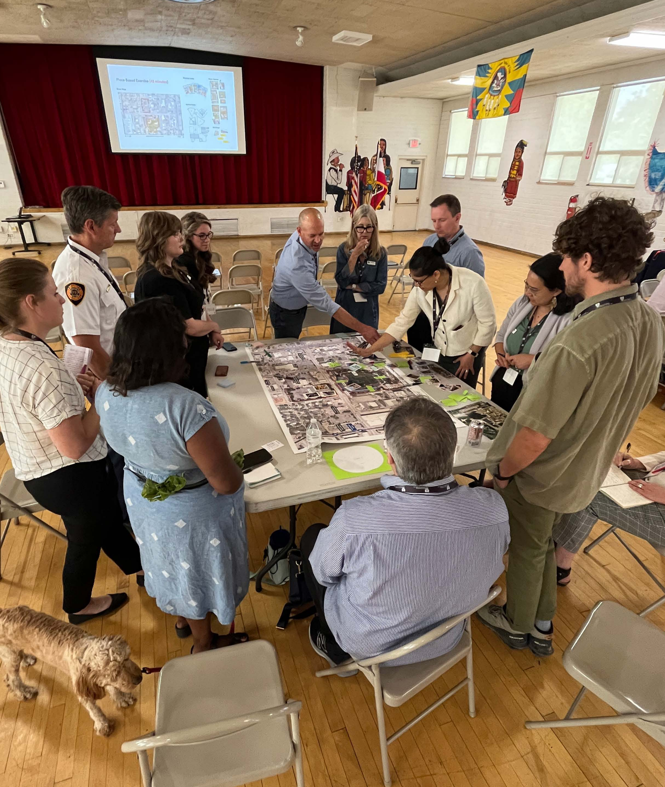 Community members gather around a site plan model during a Ballpark NEXT stakeholder ideation session; a small dog is visible in the room.