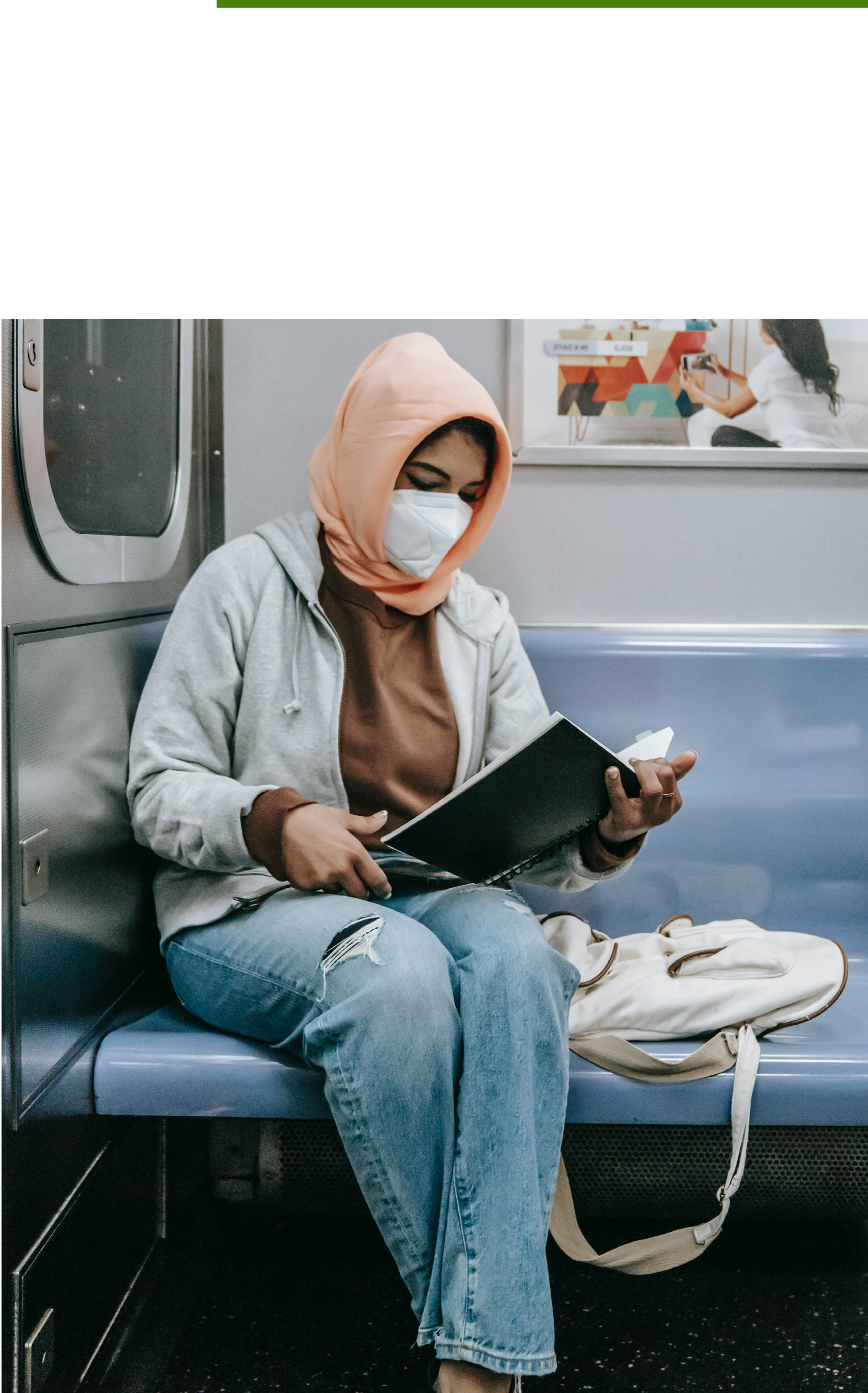 A person wearing a face mask and peach-colored hijab sits on a subway train, looking down at an open book or notebook they are holding.