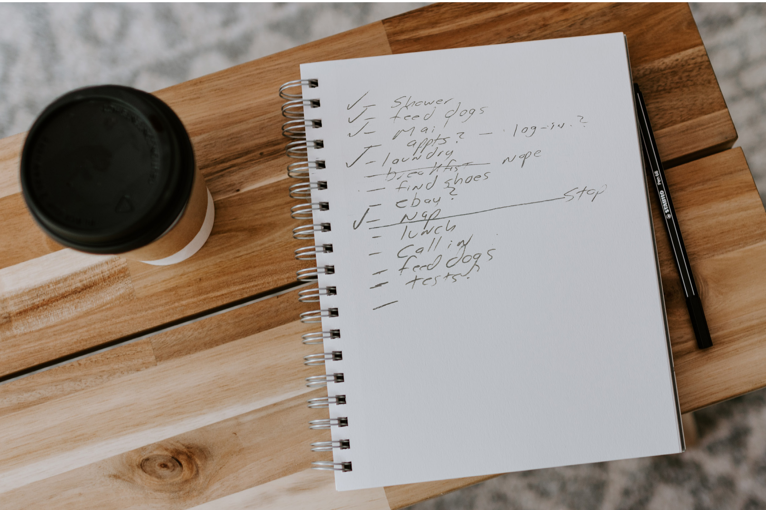 Photo of a spiral notebook on a wooden desk with a handwritten daily task list and a coffee cup.
