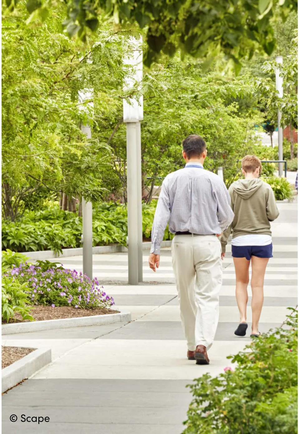 Two pedestrians walking along a paved, tree-lined walkway with lush flowering garden beds and light posts.