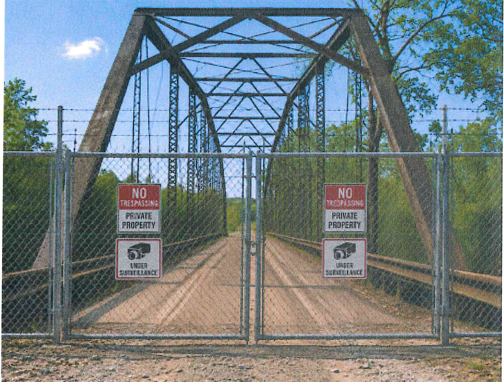 Gated entrance to the Priest Ford truss bridge with chain-link security gate and warning signs reading No Trespassing Private Property and Under Surveillance