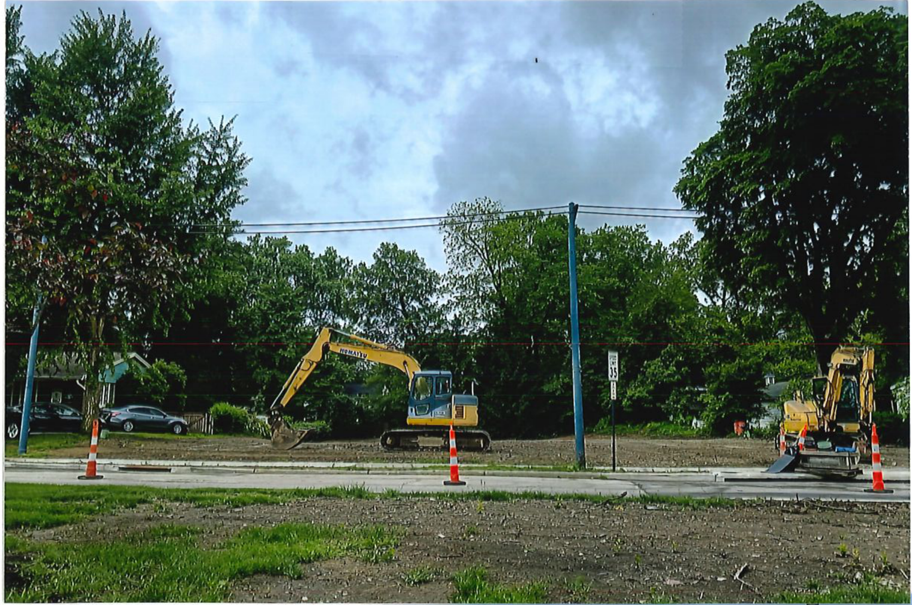 Photograph showing a construction site with heavy equipment including an excavator, traffic cones along the road, and surrounding trees.