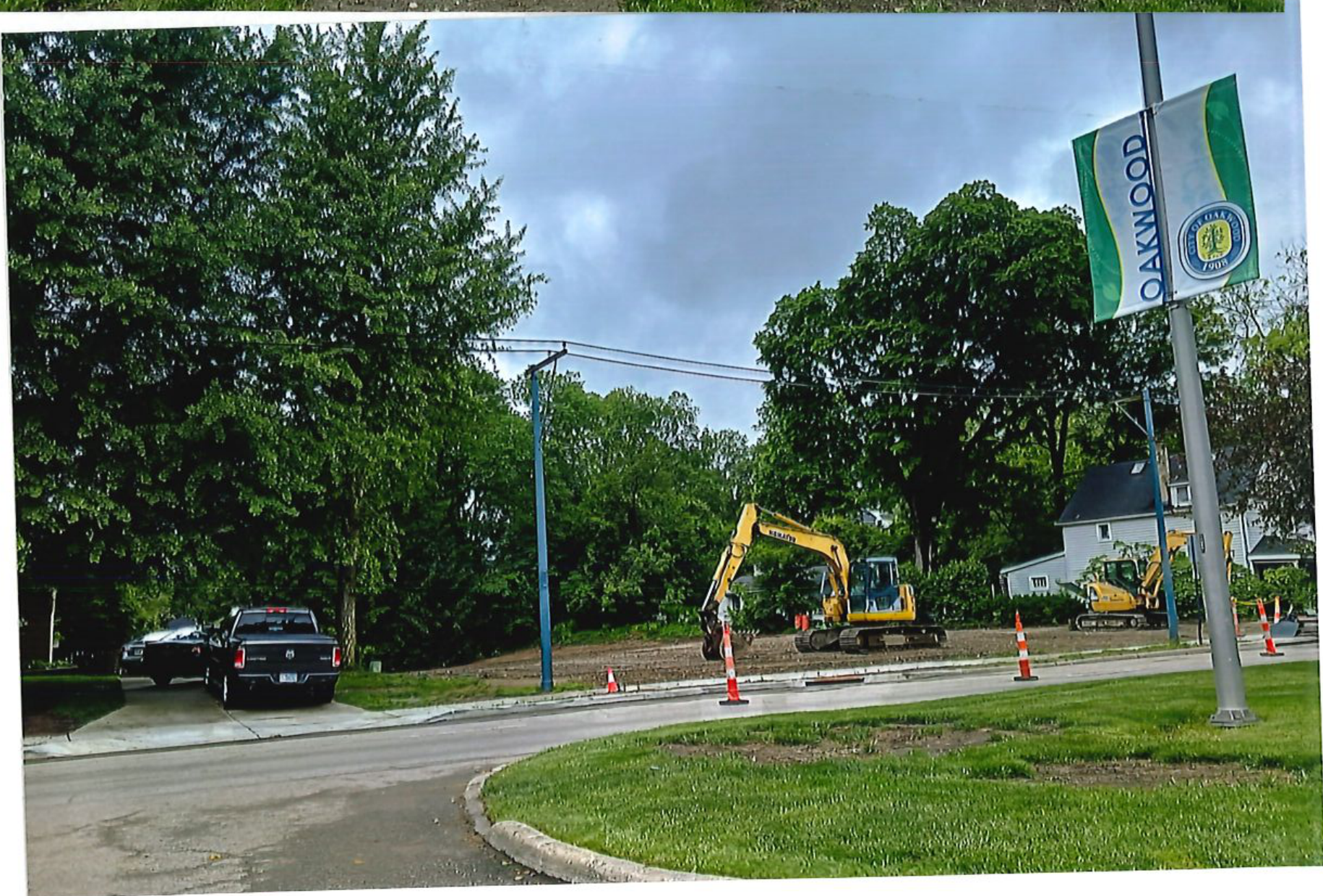 Photograph showing a street view of the Oakwood construction site with an Oakwood banner sign pole, parked vehicles, traffic cones, and an excavator.