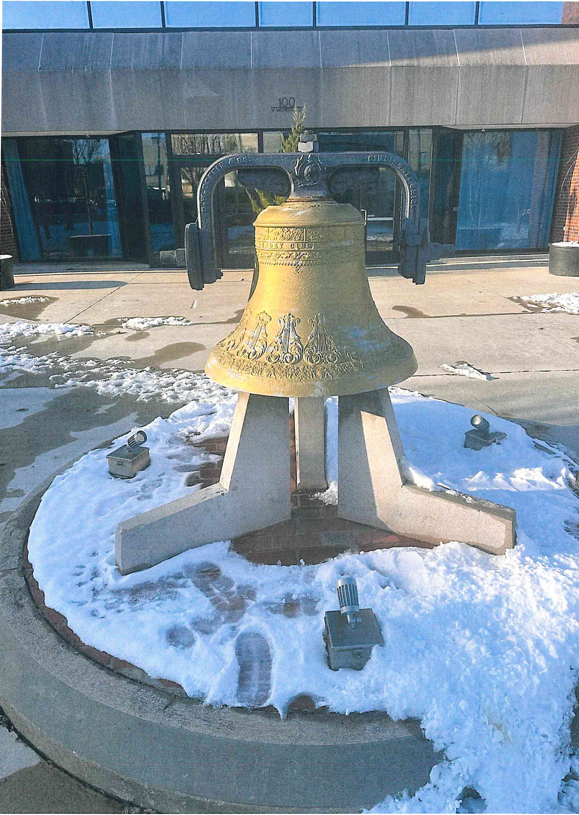 Historic bronze bell mounted on a concrete tripod pedestal outside a building, surrounded by snow on the ground