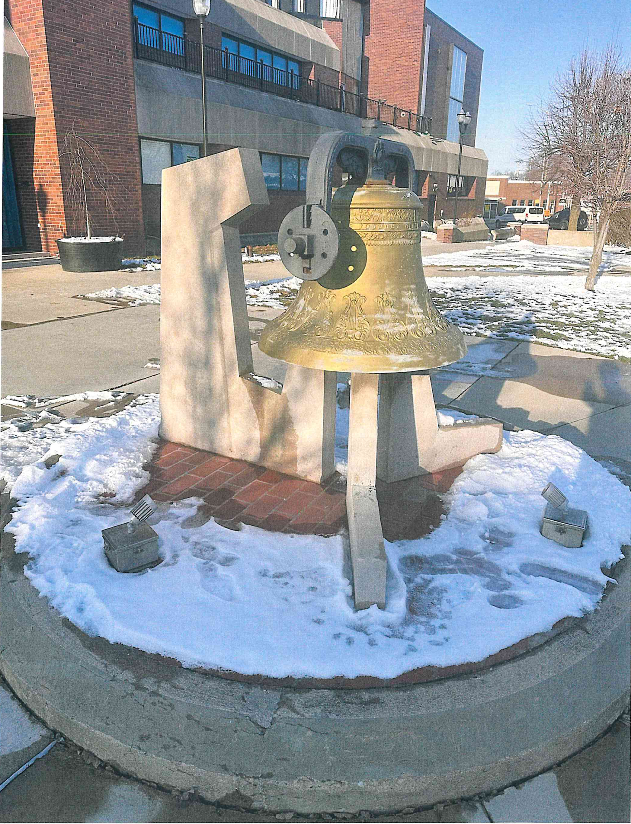Large gold bell sculpture installed on concrete pedestal outside brick building in Muncie